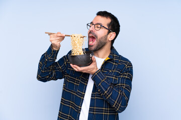 Young caucasian man over blue background holding a bowl of noodles with chopsticks and eating it