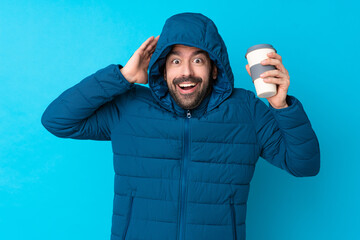 Naklejka premium Man wearing winter jacket and holding a takeaway coffee over isolated blue background with surprise expression
