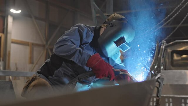 Medium long of unrecognizable worker wearing helmet and red gloves, welding metallic object in garage
