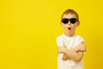 A boy in black sunglasses with his arms crossed and his mouth open funny poses on a yellow background