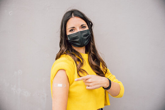 Portrait of a young woman on gray wall showing and pointing finger at medical patch after getting vaccine against Coronavirus Covid-19 infections - Millennials immunized during pandemic - Copy space