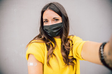 Selfie portrait of a young woman on a gray wall showing the medical patch after getting vaccinated against Coronavirus Covid-19 infections - Millennials immunized during the pandemic - Copy space