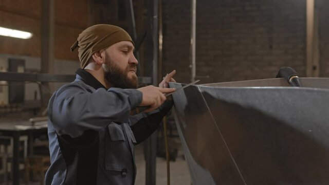 Side view waist-up of bearded man wearing worker jacket, grinding edge of boat hull with file in dockyard