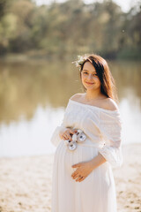 Fototapeta na wymiar pregnant woman in white dress holds little baby shoes on her stomach. Baby bump. Pregnancy and parenting. Pregnant woman with long black hair outside by a lake 
