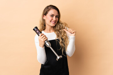 Young brazilian woman using hand blender isolated on beige background proud and self-satisfied