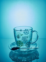 Empty mug on a saucer with a mirror reflection.In the saucer lies a spoon.The mug stands on a blue background.There are snowflakes painted on the mug 