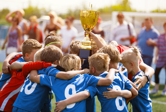 Happy Children in Football Team Winning Golden Cup in School Tournament. Young Boys in Blue Soccer Uniforms Rising Up Golden Cup. Young Coach Celebrating Success With Kids Football Players