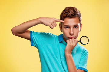 A teenage boy in a blue t-shirt with pimples pointing on his face, holding a magnifying glass....