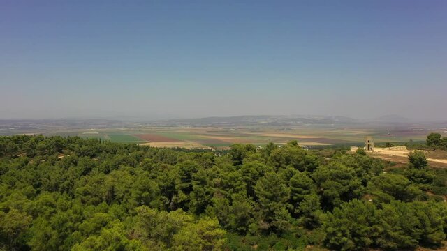 Aerial Shot Of Green Trees In Forest By Agricultural Field, Drone Flying Forward Over Landscape Against Blue Sky - Megiddo, Israel