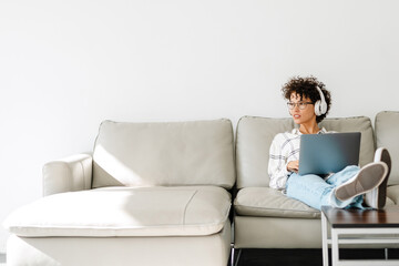 Young curly woman in headphones using laptop while sitting on couch