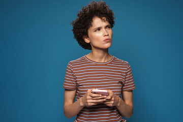 Young curly puzzled woman in striped t-shirt using mobile phone