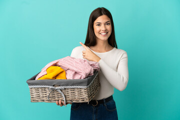 Teenager Brazilian girl holding a clothes basket isolated on blue background pointing to the side...