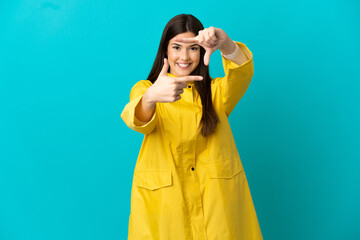 Teenager Brazilian girl wearing a rainproof coat over isolated blue background focusing face. Framing symbol