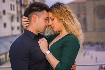 Fashionable pose of a Caucasian loving couple in a blue suit and green dress, caressing each other in the city and looking at each other about to kiss