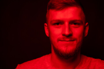 Young attractive male person posing in contrast red light. Portrait of smiling handsome guy in dark studio with red neon lightning. Nice man looks friendly at camera, fashion beauty concept