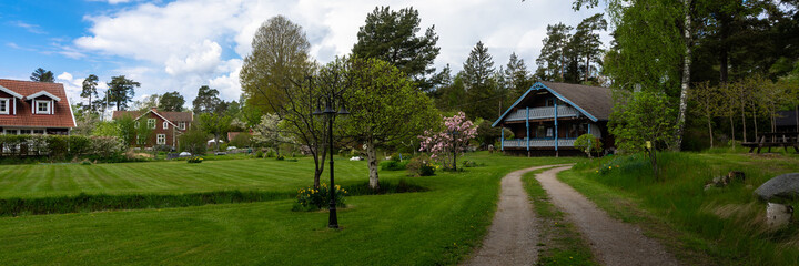 Panorama of countryside. Old two story wooden house by the road.Villa cottage made of dark wood with large terrace and balcony. Garden design with flowering trees and daffodils. Green lawn is mowed.