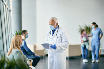 Senior doctor with face mask reading medical report while communicating with female patient in...