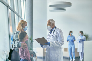 Mature pediatrician talking to mother and daughter in hallway at medical clinic during COVID-19 pandemic.