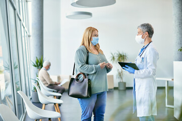 Doctor with face mask using touchpad while talking to female patient at hospital hallway.