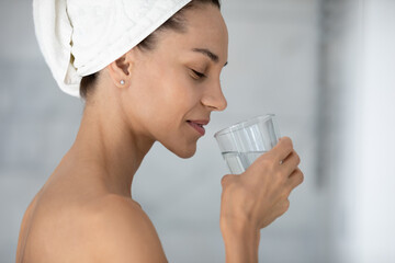 Happy woman wrapped in towel enjoying high quality fresh pure water, holding transparent glass, drinking after morning bath for skincare and healthy diet. Aqua balance, hydration concept