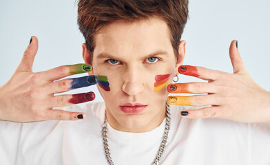 Young gay man is standing in the studio and posing for a camera. Painted by multi colored makeup