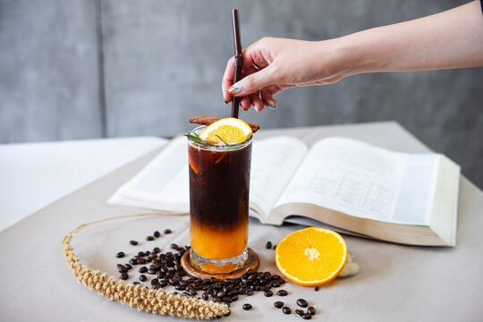 Orange black coffee and open book on the coffee table surrounded with coffee bean, hand touching a straw.