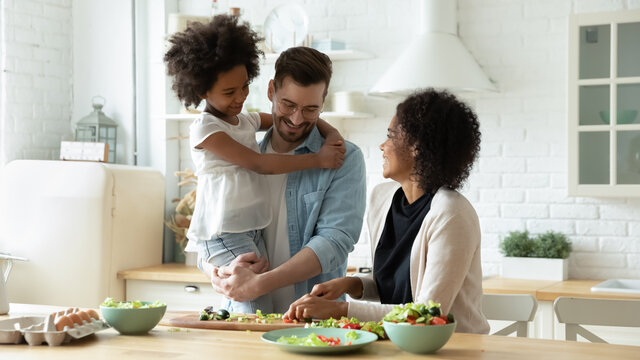 Smiling Young Multiracial Mom And Dad Cook Healthy Delicious Vegetarian Salad With Ethnic Small Daughter. Happy Multiethnic Family With Girl Child Have Fun Prepare Food In Kitchen. Adoption Concept.