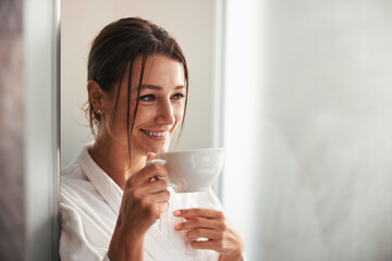 Laughing beautiful elegant lady in white bathrobe looking away while holding cup in hand in room indoors - obrazy, fototapety, plakaty