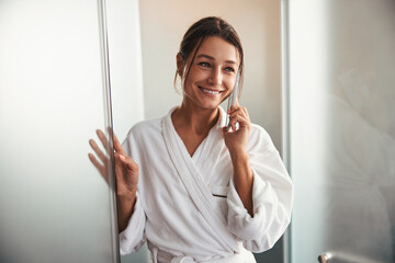Brunette Caucasian charming business woman in white bathrobe having dialogue on cell phone in...