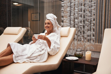 Beautiful adorable Caucasian woman with towel on her head relaxing and drinking herbal tea in...