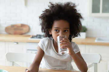 Thirsty small African American girl child sit at home kitchen feel dehydrated enjoy clean clear pure mineral water from glass. Little teen ethnic kid sip aqua for body refreshment. Hydration concept. - obrazy, fototapety, plakaty