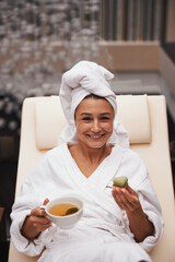 Charming elegant Caucasian woman holding cup of tea and green cake i hands while looking at the photo camera in wellness center