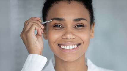 Face portrait of happy Black millennial girl plucking brows with tweezers smiling at camera despite painful procedures. Young woman tweezing eyebrows and correcting shapes at home. Close up - obrazy, fototapety, plakaty