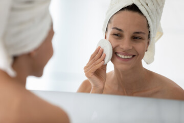 Happy beautiful girl cleansing facial skin with skincare exfoliating sponge, washing face with cleanser, lotion for pure pores after shower. Home cosmetic procedures, acne treatment concept