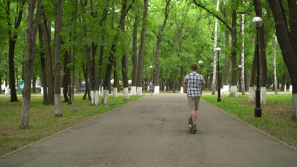 A young European man in a shirt and shorts rides an electric scooter in the park.