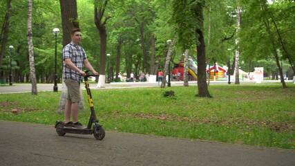 A young European man in a shirt and shorts rides an electric scooter in the park.