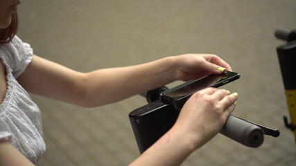 Obraz premium A young woman finishes a ride on a sharing e-scooter. The girl took the phone off the scooter. Steering wheel close-up.