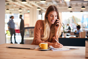 Young Businesswoman Sitting At Desk On Phone Call In Modern Open Plan Office