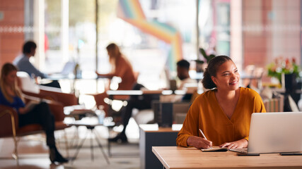 Young Businesswoman Sitting At Desk Writing In Notebook In Modern Open Plan Office
