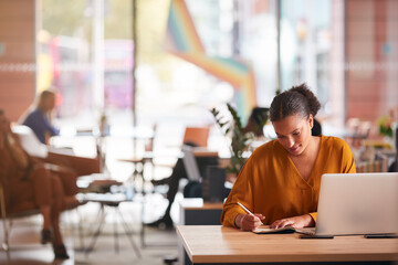 Young Businesswoman Sitting At Desk Writing In Notebook In Modern Open Plan Office