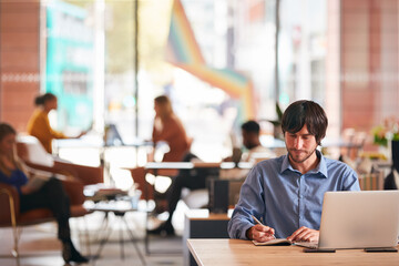 Businessman Sitting At Desk Writing In Notebook In Modern Open Plan Office