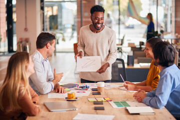 Businessman Giving Presentation To Colleagues Sitting Around Table In Modern Open Plan Office