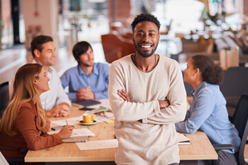 Portrait Of Businessman With Colleagues In Background Sitting Around Table In Open Plan Office