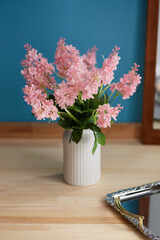 Still life photo of white ceramic vase with bunch of pink phlox flowers against blue wall. Scandinavian bowl with ribbed surface is located on wooden table near silver server and mirror in frame.  