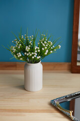 Still life photo of white ceramic vase with bunch of white gypsophila flowers against blue wall. Scandinavian bowl with ribbed surface is located on wooden table near silver server and mirror.  