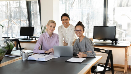 Portrait of three confident diverse businesswomen sitting at table with laptop in office, smiling successful colleagues employees involved in teamwork, looking at camera, posing for corporate photo