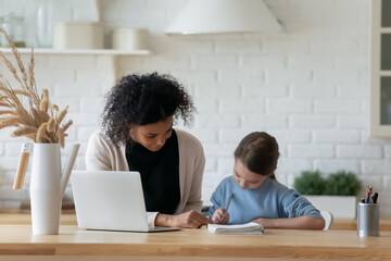 Loving young African American mom study online on laptop do homework with Caucasian little daughter at home. Caring biracial mother help small girl child with school preparation. Diversity concept.