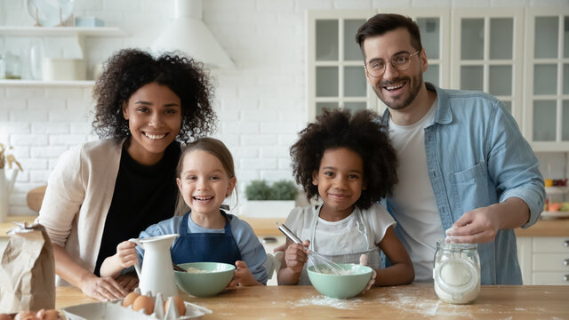 Portrait of smiling young multiethnic family with diverse children have fun cooking together at home kitchen. Happy multiracial mom and dad with daughters bake pastry with dough. Adoption concept.