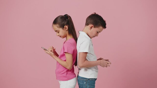 Little boy and girl standing back to back and networking on smartphones, curious sister peeking into brothers phone