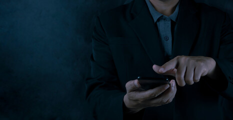 A businessman in a black suit is touching his smartphone on a black background.